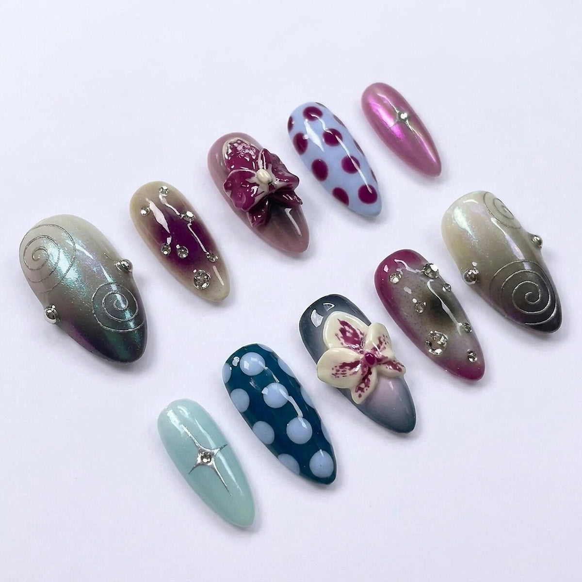 A set of ten almond-shaped press-on nails featuring diverse designs including purple and white polka dots, iridescent finishes, silver swirl patterns, small clear rhinestones, and three-dimensional orchid flower embellishments.