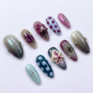 A set of ten almond-shaped press-on nails featuring diverse designs including purple and white polka dots, iridescent finishes, silver swirl patterns, small clear rhinestones, and three-dimensional orchid flower embellishments.
