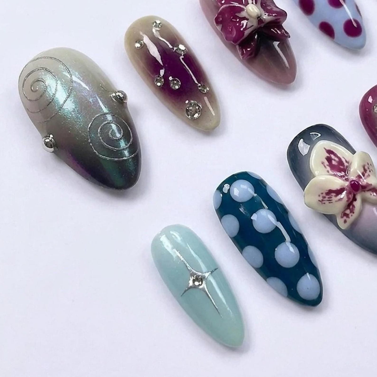 A collection of almond-shaped press-on nails featuring diverse designs including iridescent swirls, embedded rhinestones, 3D floral accents, light blue polka dots on a dark teal base, and a pale blue nail with a silver star emblem.