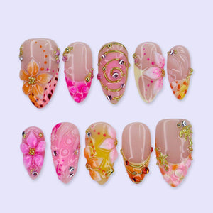 A set of ten press-on nails featuring a nude base with intricate 3D designs including orange and pink floral motifs, gold wire swirls, metallic studs, rhinestones, and small gold charms shaped like palm trees and starfish.