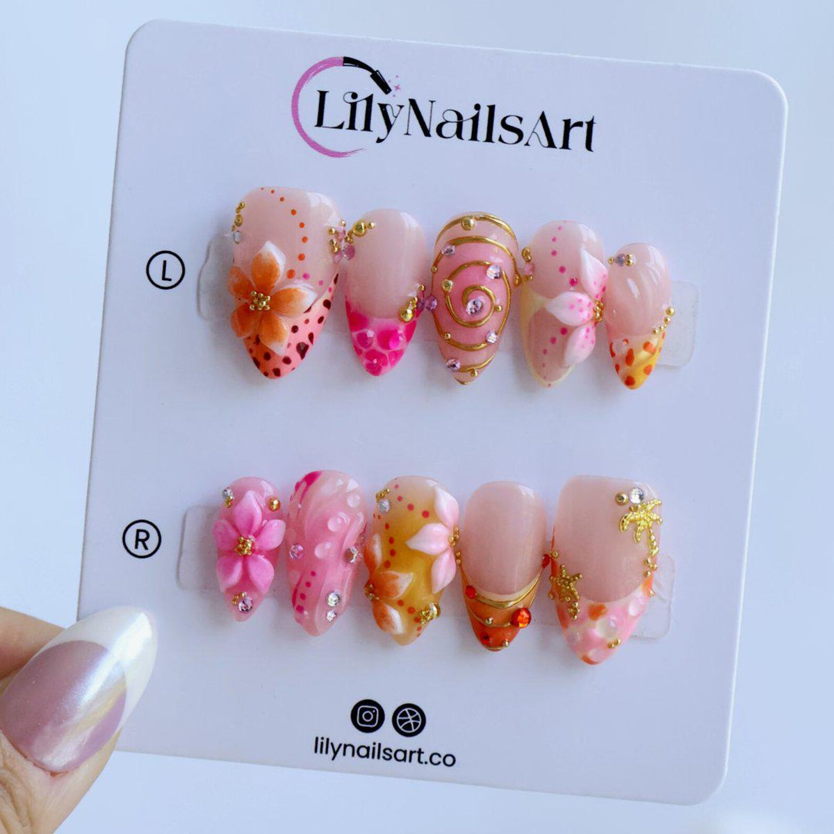 A set of ten press-on nails on a white display card labeled LilyNailsArt. The nails feature a translucent base with pink, orange, and gold accents, including 3D floral designs, gold swirls, small rhinestones, and a gold palm tree charm.