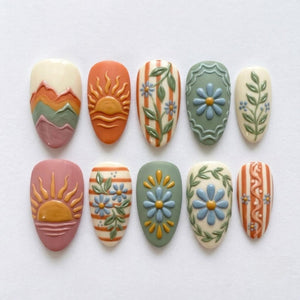 A set of ten almond-shaped press-on nails featuring embossed designs including layered mountain ranges, rising suns over water, vertical stripes with floral vines, daisies within scalloped frames, and intricate botanical patterns in muted tones.