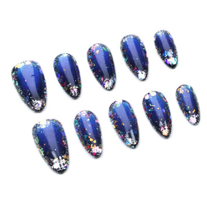 A set of ten almond-shaped press-on nails featuring a deep navy blue base color. Each nail is decorated with multi-colored iridescent foil flakes and glitter particles that create a reflective, galaxy-like appearance across the surface.