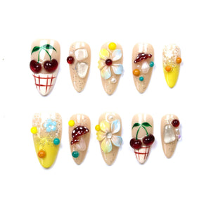 A set of ten almond-shaped press-on nails featuring various 3D embellishments including red cherries, white and brown mushrooms, multi-colored floral designs, clear crystals, and yellow accents on a nude, glitter-flecked base.