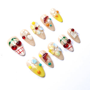 A set of ten almond-shaped press-on nails featuring a nude base with various 3D embellishments including red cherries, white and yellow flowers, small mushrooms, and colorful beads. Some nails have red grid patterns or yellow tips.