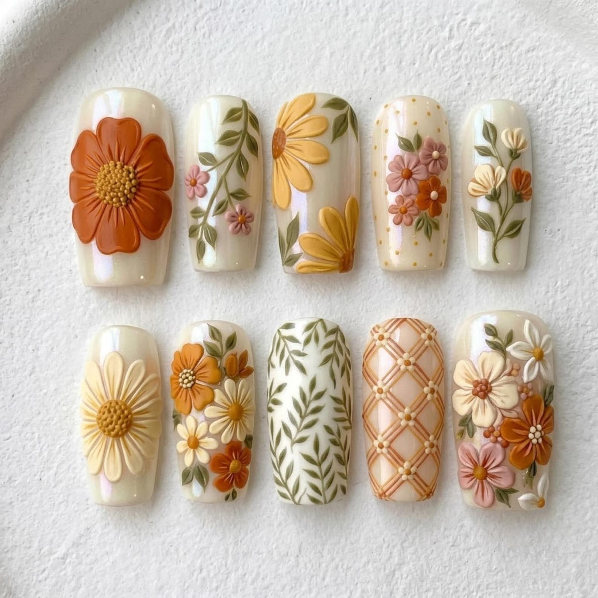 A set of ten press-on nails featuring raised 3D floral designs in shades of orange, yellow, cream, and green. The patterns include individual large flowers, delicate leafy vines, small clusters of blossoms, and a geometric lattice grid.