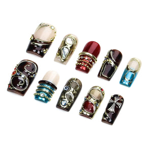 A set of ten press-on nails featuring various designs including deep red, brown, and blue glossy finishes. The nails are decorated with gold-toned metal accents, small embedded rhinestones, and a silver-toned cross emblem.