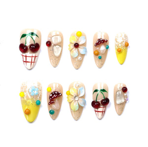 A set of ten almond-shaped press-on nails featuring various 3D embellishments including red cherries, white and red mushrooms, multi-colored flowers, clear crystals, yellow gradients, and intricate lace patterns on a nude base.