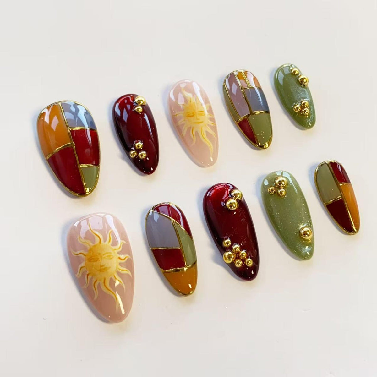 A set of ten almond-shaped press-on nails featuring a variety of designs including deep red, olive green, and orange color-blocked patterns with gold lines, sun illustrations on light pink bases, and small gold metallic bead accents.