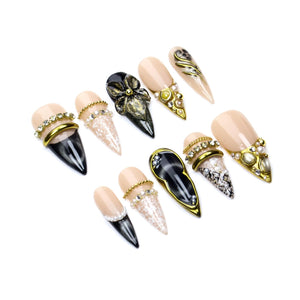 A set of ten stiletto-shaped press-on nails featuring a nude base with black accents. The designs include gold metallic bands, clear rhinestones, white lace patterns, pearl embellishments, and a three-dimensional black floral motif.