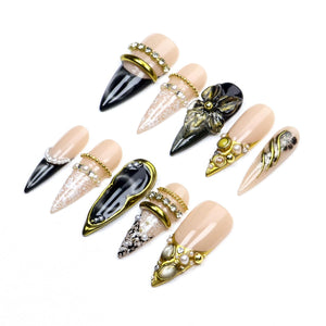A set of ten stiletto-shaped press-on nails featuring a nude base with black, gold, and white accents. The designs include intricate lace patterns, metallic gold bands, small white pearls, and a three-dimensional black floral embellishment.