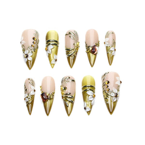 A set of ten stiletto-shaped press-on nails featuring a gradient of nude and olive green. The nails are decorated with raised white floral accents, metallic gold swirls, and small amber-colored rhinestones arranged in various patterns.