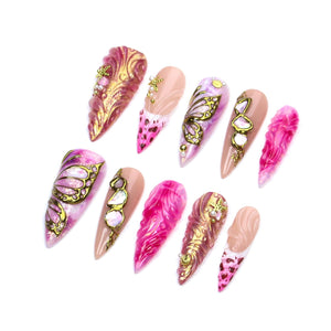 A set of ten stiletto-shaped press-on nails featuring a variety of designs including pink and gold marbled patterns, gold butterfly wing accents, iridescent stone embellishments, and small gold star charms on a light pink base.