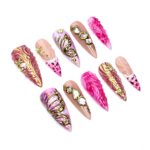 A set of ten stiletto-shaped press-on nails featuring a variety of designs in shades of pink, gold, and nude. The nails are decorated with raised gold butterfly wing patterns, textured swirls, small starfish charms, and embedded rhinestones.