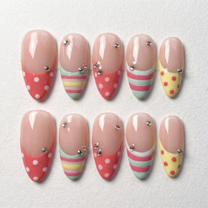 A set of ten almond-shaped press-on nails featuring a nude base with colorful tips. The tips display patterns of red polka dots, horizontal stripes in pink, yellow, and mint green, and yellow polka dots, all accented with small clear rhinestones.