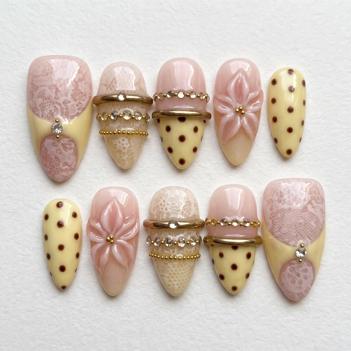 A set of ten press-on nails featuring a mix of designs including pale pink lace patterns, cream-colored bases with brown polka dots, raised pink floral accents, and horizontal gold-toned metallic bands embellished with small clear rhinestones.