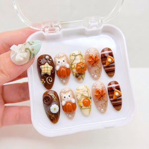 A set of ten press-on nails in a white plastic case, featuring autumn-themed 3D designs including orange pumpkins, white kittens, maple leaves, plaid patterns, brown stripes, small buttons, and miniature pie slices with cream dollops.