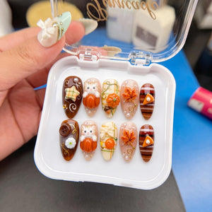 A set of ten press-on nails in a clear plastic case, featuring autumn-themed designs including 3D cats in pumpkins, orange maple leaves, plaid patterns, brown stripes, small white flowers, buttons, and miniature pie slices with pearls.