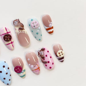 A set of ten almond-shaped press-on nails featuring various designs including pink and white plaid patterns, light blue and pink polka dots, brown cat figures, small white pearls, and raised circular button embellishments in brown and cream.