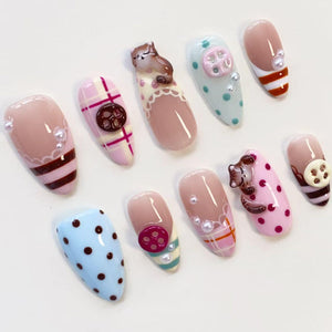 A set of ten almond-shaped press-on nails featuring various designs including pink and white plaid, light blue and pink polka dots, brown cat figures, and small buttons. The nails are decorated with white pearls and glossy finishes.
