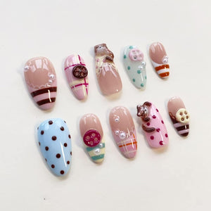 A set of ten press-on nails featuring various designs including pink and blue polka dots, plaid patterns, brown stripes, small white pearls, and three-dimensional sculpted brown bear figures and circular buttons attached to the nail surfaces.