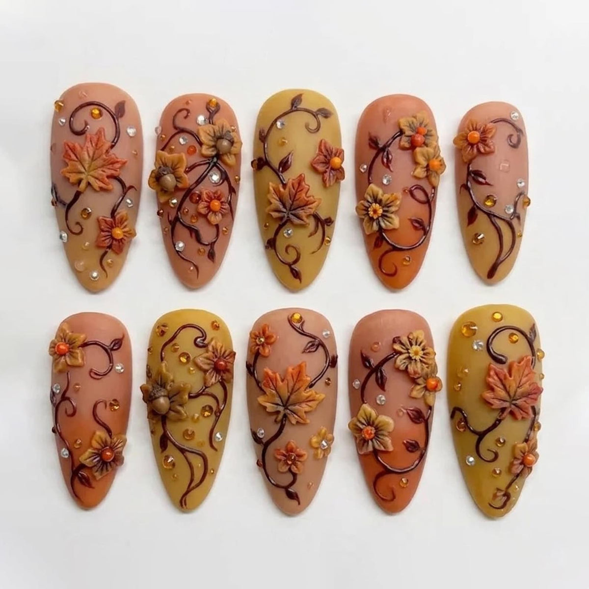 A set of ten almond-shaped press-on nails featuring a warm autumn color palette of tan, mustard yellow, and burnt orange. Each nail is decorated with raised 3D maple leaves, small flowers, dark brown swirling vines, and tiny clear rhinestones.