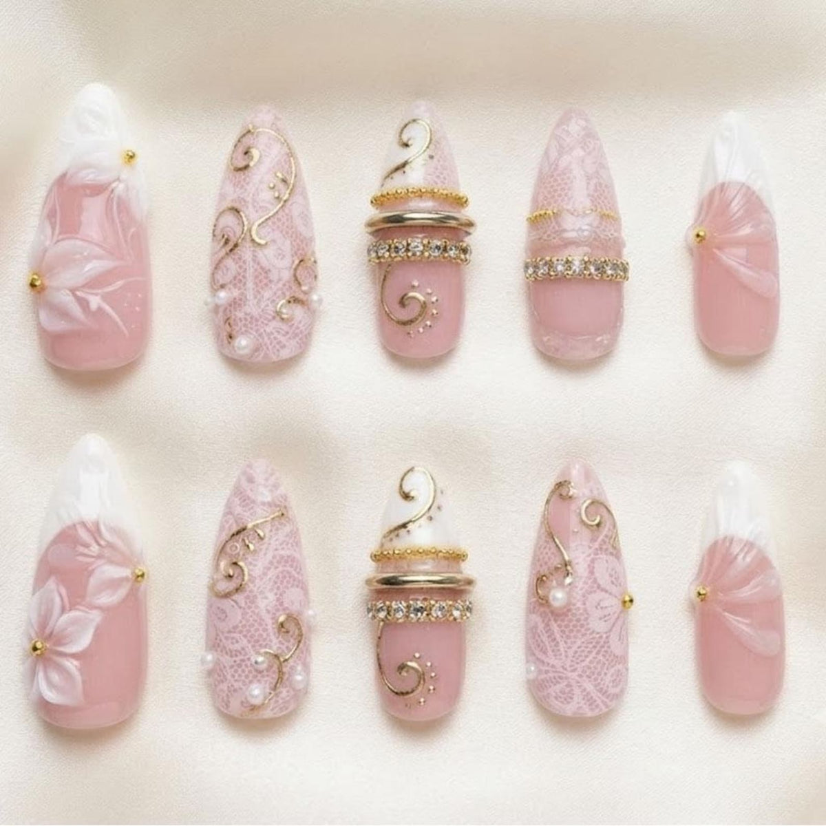 A set of ten almond-shaped press-on nails in shades of soft pink and white. The designs feature intricate white lace patterns, raised floral accents, gold metallic swirls, small pearls, and horizontal bands of clear rhinestones.