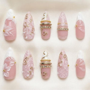 A set of ten almond-shaped press-on nails in shades of soft pink and white. The designs feature intricate white lace patterns, raised floral accents, gold metallic swirls, small pearls, and horizontal bands of clear rhinestones.