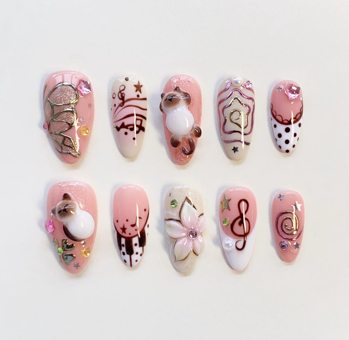 A set of ten press-on nails featuring a pink and white color palette with various 3D embellishments including miniature cats, a flower, musical notes, treble clefs, stars, polka dots, and small rhinestones arranged in a decorative pattern.