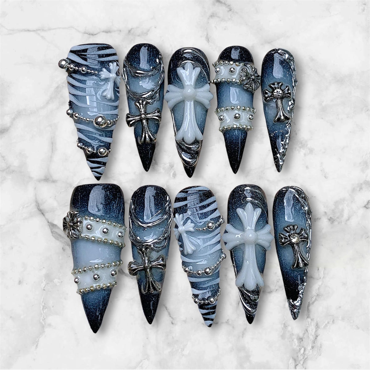 A set of ten stiletto-shaped press-on nails featuring a dark blue and black gradient base. The designs include white zebra stripes, silver metallic chains, small silver studs, and raised white or silver cross emblems on each nail.