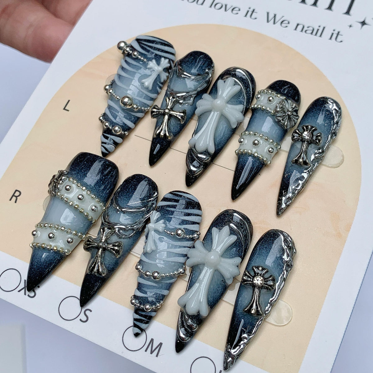 A set of ten stiletto-shaped press-on nails featuring a dark blue to light blue gradient base. The nails are decorated with silver metallic chains, small silver studs, and raised white cross emblems. The nails are displayed on a card.