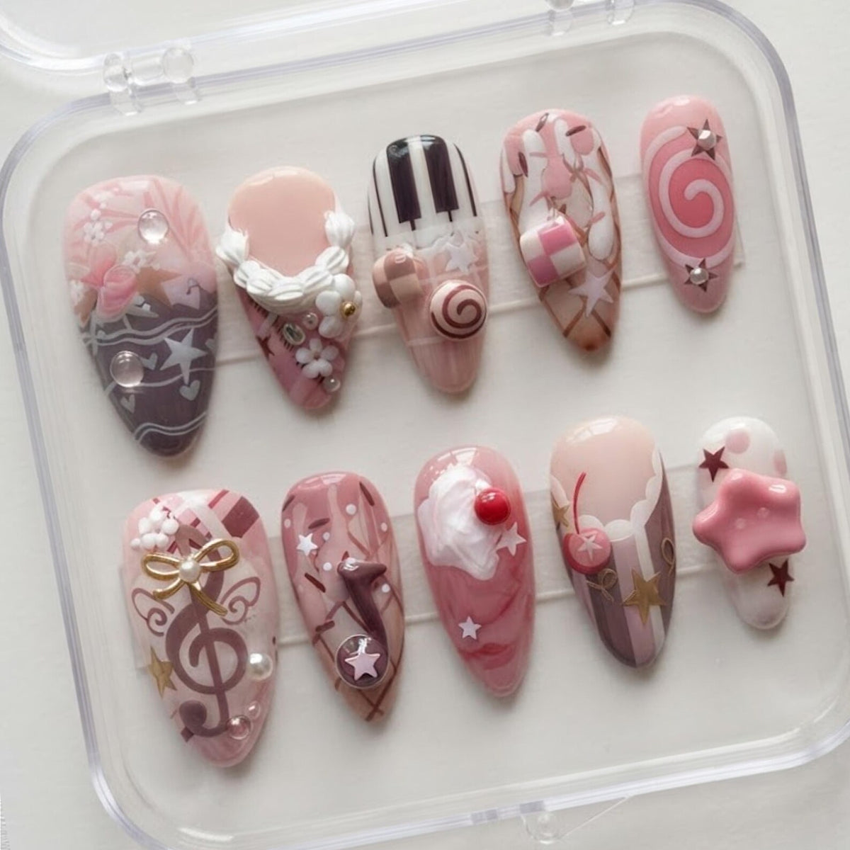 A set of ten almond-shaped press-on nails in a clear plastic case. The nails feature pink, white, and brown color palettes with 3D embellishments including white cream swirls, a cherry, a gold bow, stars, and piano key patterns.
