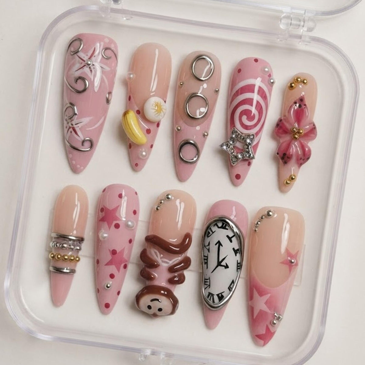 A set of ten almond-shaped press-on nails in a clear plastic case. The nails feature a pink and nude color palette with various 3D embellishments, including silver rings, a yellow banana, a star, a monkey figure, and a melting clock.