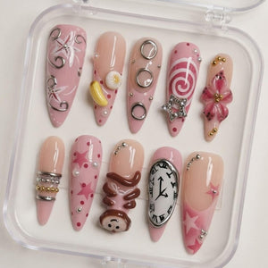 A set of ten almond-shaped press-on nails in a clear plastic case. The nails feature a pink and nude color palette with various 3D embellishments, including silver rings, a yellow banana, a star, a monkey figure, and a melting clock.