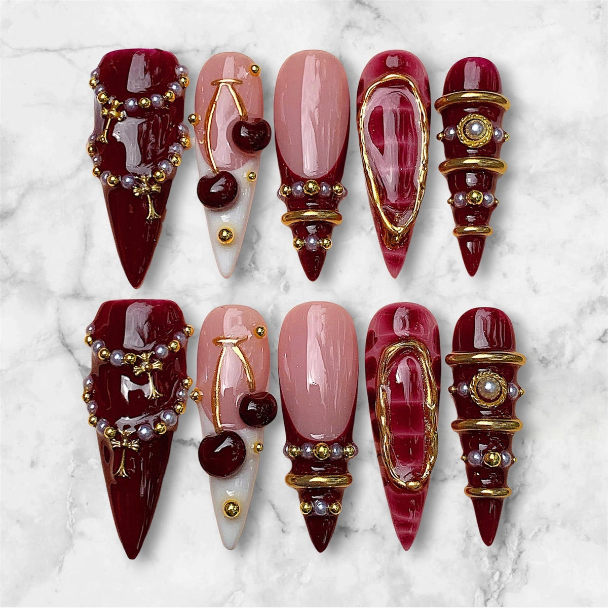 A set of ten press-on nails featuring a stiletto shape. The designs include deep burgundy polish with gold chains and pearls, light pink bases with dark red cherry charms, French tips, and burgundy nails with gold rings and studs.