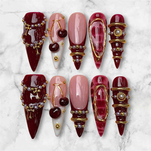 A set of ten press-on nails featuring a stiletto shape. The designs include deep burgundy polish with gold chains and pearls, light pink bases with dark red cherry charms, French tips, and burgundy nails with gold rings and studs.