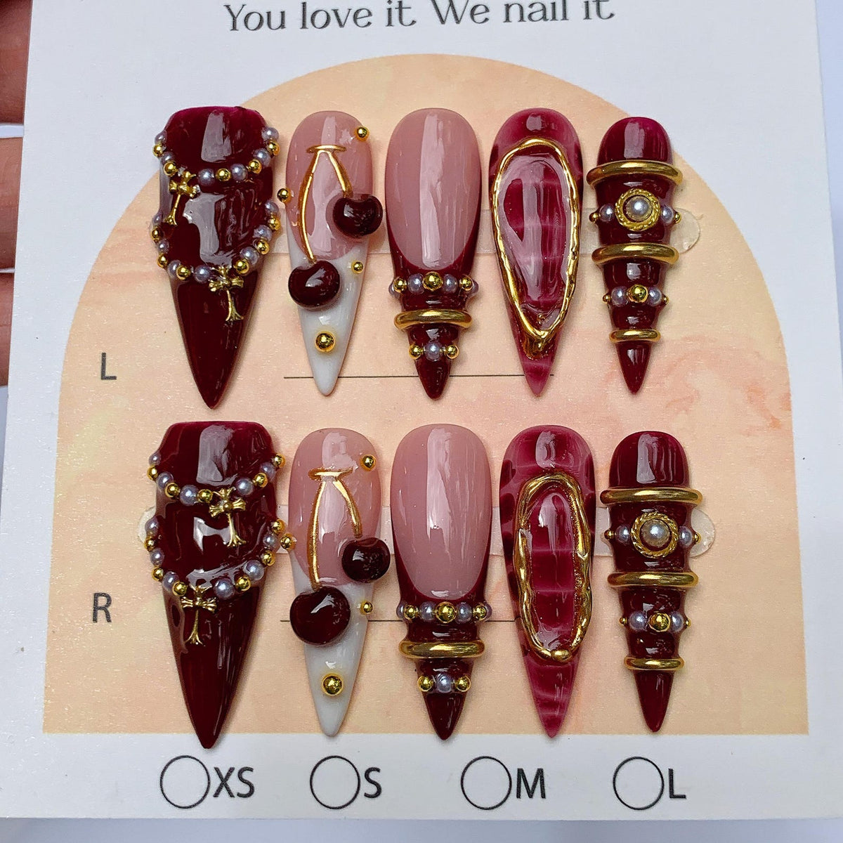 A set of ten press-on nails in a stiletto shape, featuring deep burgundy and nude tones. The designs include gold-toned charms, small white pearls, cherry motifs, and metallic gold accents. The display card includes the text You love it. We nail it.
