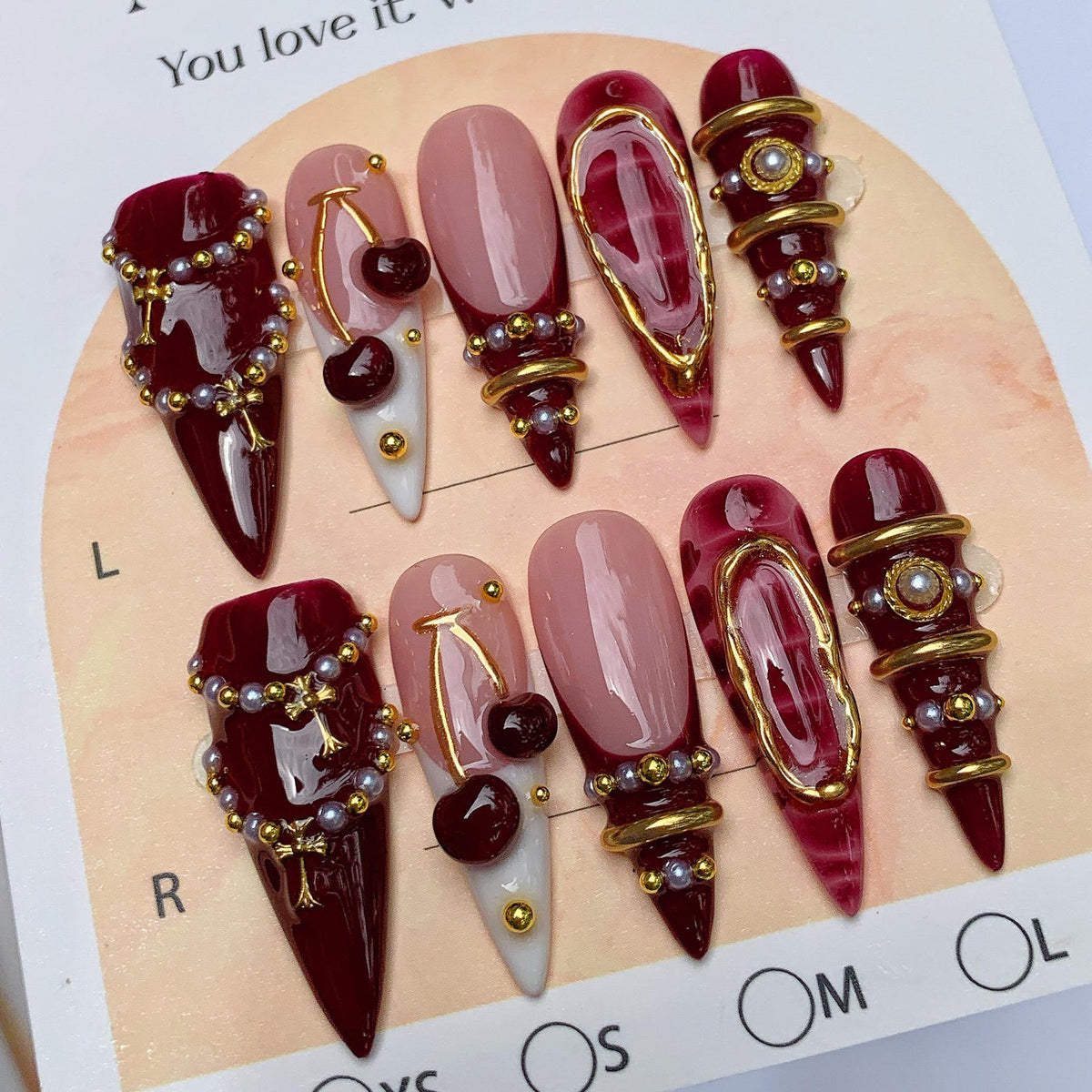 A set of ten press-on nails featuring a deep burgundy and nude color palette. The designs include glossy burgundy tips, cherry charms, gold metallic rings, and small pearl accents. The nails are arranged on a display card labeled L and R.