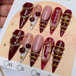 A set of ten stiletto-shaped press-on nails featuring a deep burgundy and nude color palette. The designs include gold cross charms, small pearl accents, cherry motifs, and gold metallic rings. The nails are displayed on a card labeled L, R, XS, S, a