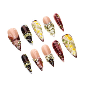 A set of ten stiletto-shaped press-on nails featuring a mix of designs including nude bases with 3D floral accents, deep burgundy tortoiseshell patterns, gold metallic bands, small clear rhinestones, and yellow and white floral motifs.