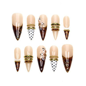 A set of ten stiletto-shaped press-on nails featuring a nude base with various designs including brown tortoiseshell patterns, black polka dots on white, gold-toned metallic bands, small rhinestones, and raised floral embellishments.