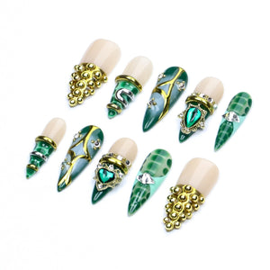 A set of ten press-on nails featuring a mix of nude and deep green colors. The designs include gold metallic studs, intricate gold filigree patterns, clear rhinestones, and a central green heart-shaped gem set in a gold frame.