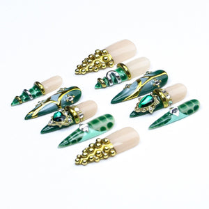 A set of ten press-on nails featuring a stiletto shape with a nude base and various green accents. Designs include gold metallic studs, silver and gold foil detailing, emerald-colored rhinestones, and a green crocodile skin pattern.