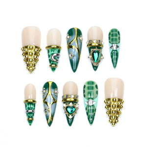 A set of ten press-on nails featuring a mix of nude and deep green tones. The designs include gold metallic studs, intricate gold filigree patterns, emerald-colored gemstone accents, and textured green scales on various pointed shapes.