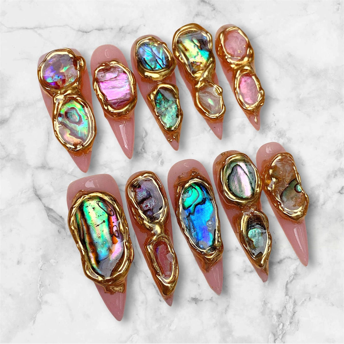 A set of ten stiletto-shaped press-on nails with a light pink base. Each nail features irregular, iridescent abalone shell fragments encased in raised, textured gold metallic borders that follow the organic shapes of the shells.