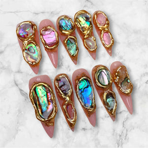 A set of ten stiletto-shaped press-on nails with a light pink base. Each nail features irregular, iridescent abalone shell fragments encased in raised, textured gold metallic borders that follow the organic shapes of the shells.