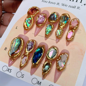 A set of ten press-on nails with a light pink base, each featuring irregular, iridescent abalone shell fragments encased in raised, textured gold-colored borders. The nails are arranged on a display card labeled with sizes.