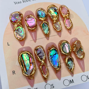 A set of ten stiletto-shaped press-on nails in a pale pink base color. Each nail features irregular, iridescent abalone shell fragments encased in textured gold-toned metal borders. The nails are arranged on a display card labeled L and R.