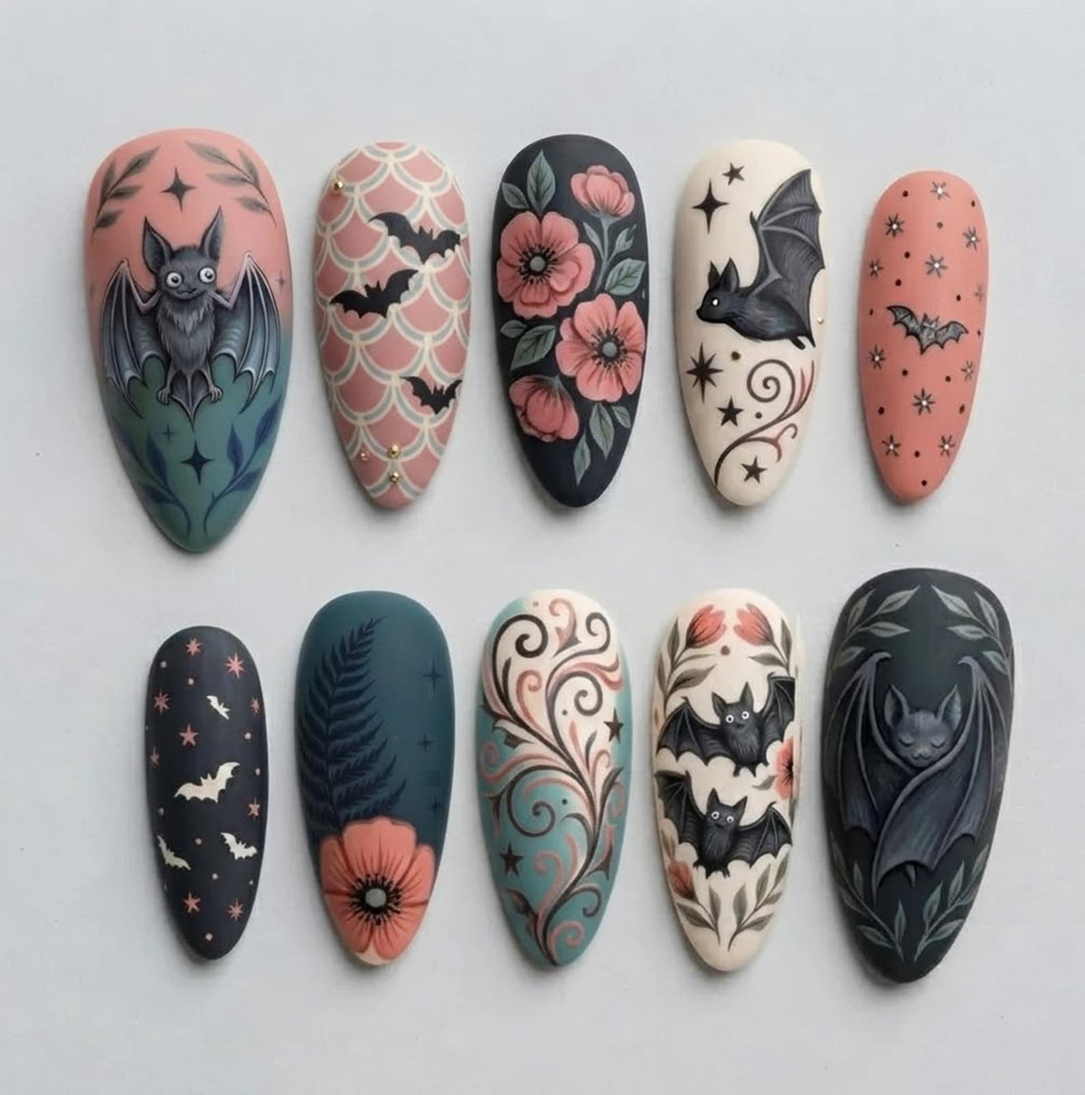 A set of ten almond-shaped press-on nails featuring intricate hand-painted designs. The collection includes motifs of bats, pink flowers, fern leaves, swirling filigree patterns, and stars set against backgrounds of black, cream, and dusty rose.