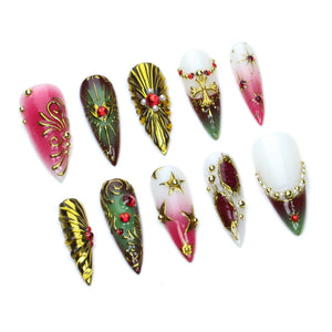 A set of ten stiletto-shaped press-on nails featuring a variety of designs in red, green, white, and gold. The nails are decorated with metallic gold filigree, red rhinestones, gold cross charms, star motifs, and small white pearls.